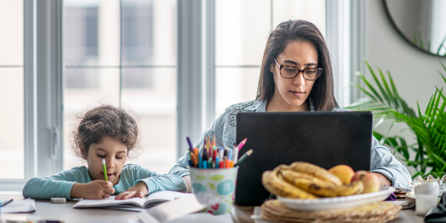 A woman wearing glasses works on a laptop at a table, while a young child sits beside her, focused on drawing or writing in a notebook, with fruit and art supplies nearby