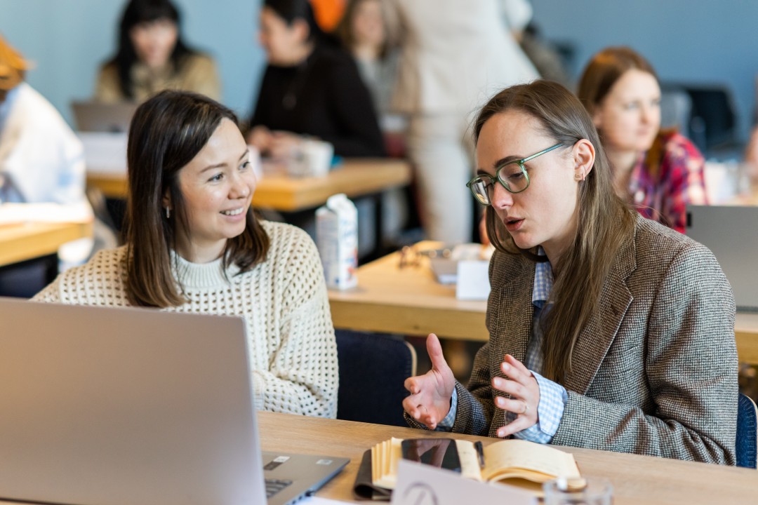 A woman in glasses is gesturing while talking to another woman sitting beside a laptop