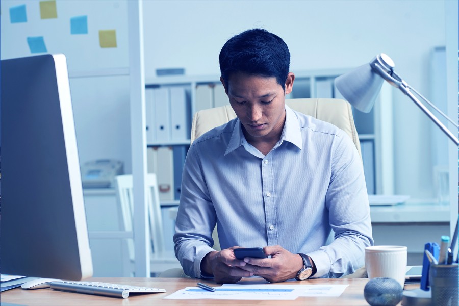 A man in a light blue shirt is sitting at his office desk using a smartphone