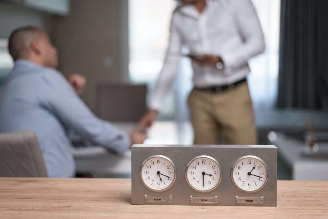A metal desk clock showing three international time zones sits on a wooden table