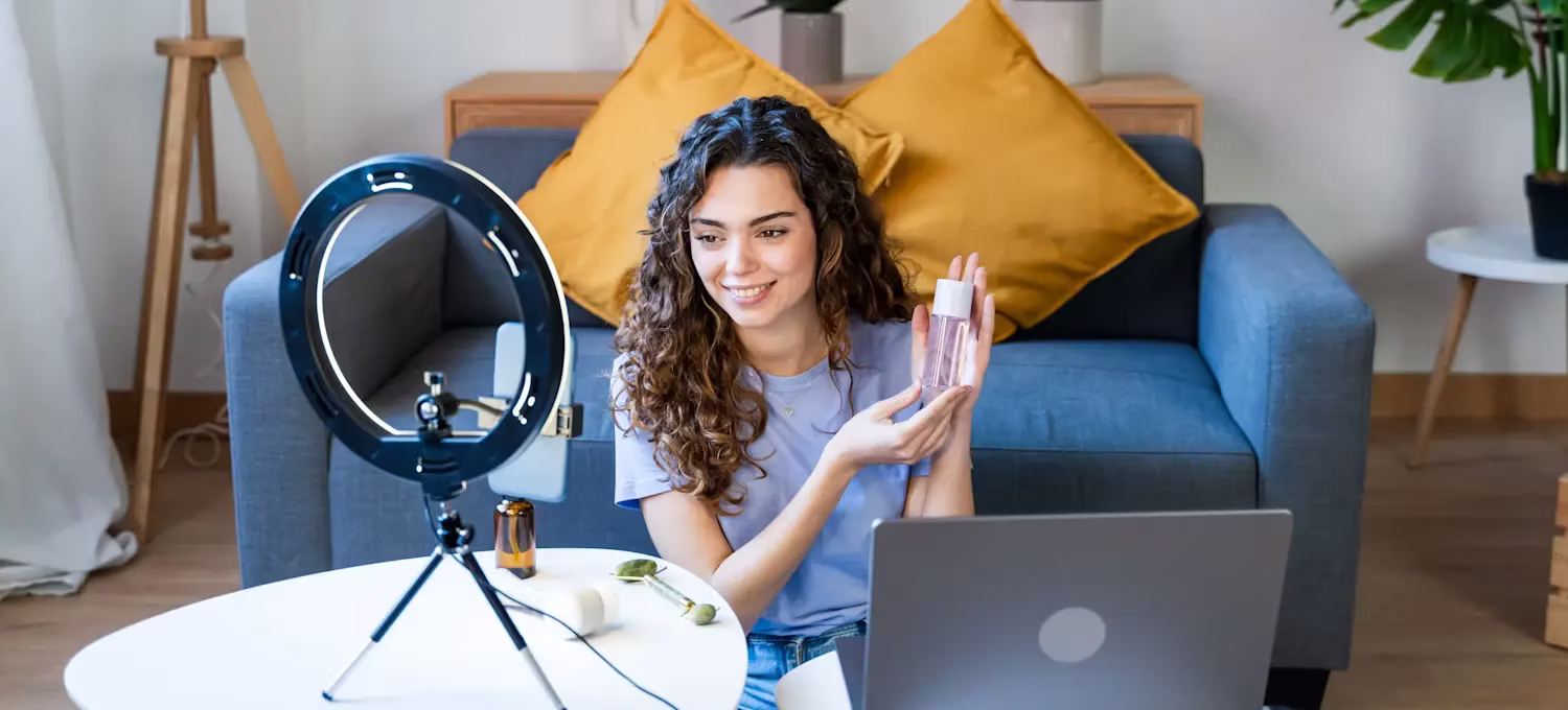 A female influencer filming a beauty product review using a ring light, smartphone, and laptop in a bright living room