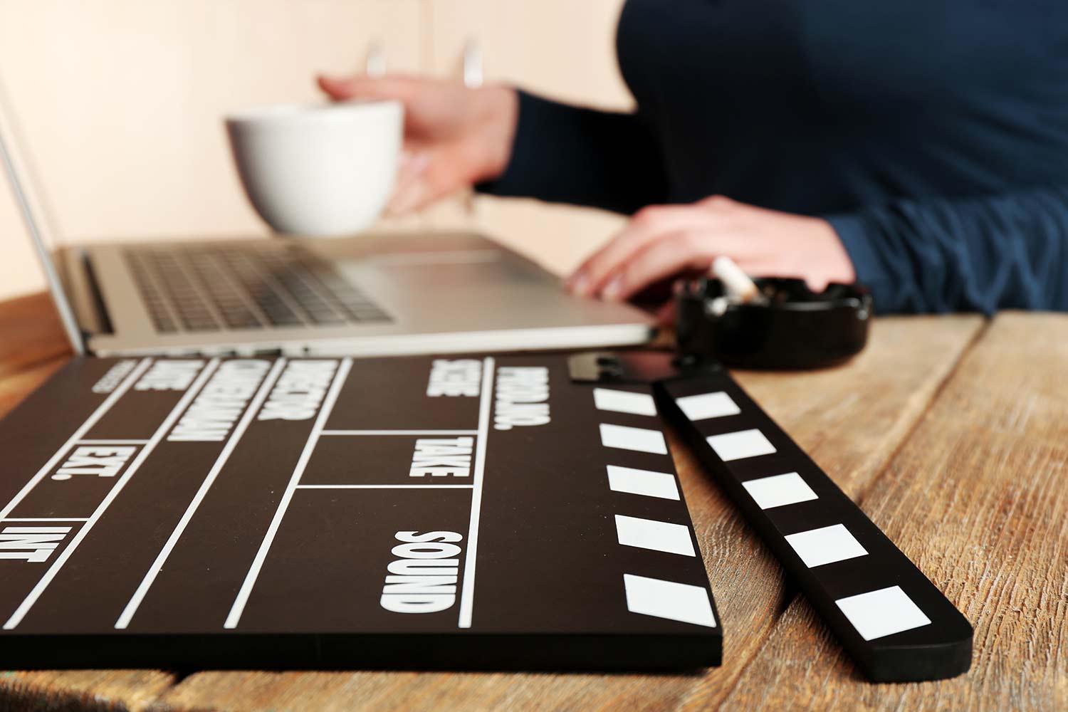 Close-up of a movie clapperboard on a wooden desk next to a screenwriter using a laptop and holding a white coffee cup