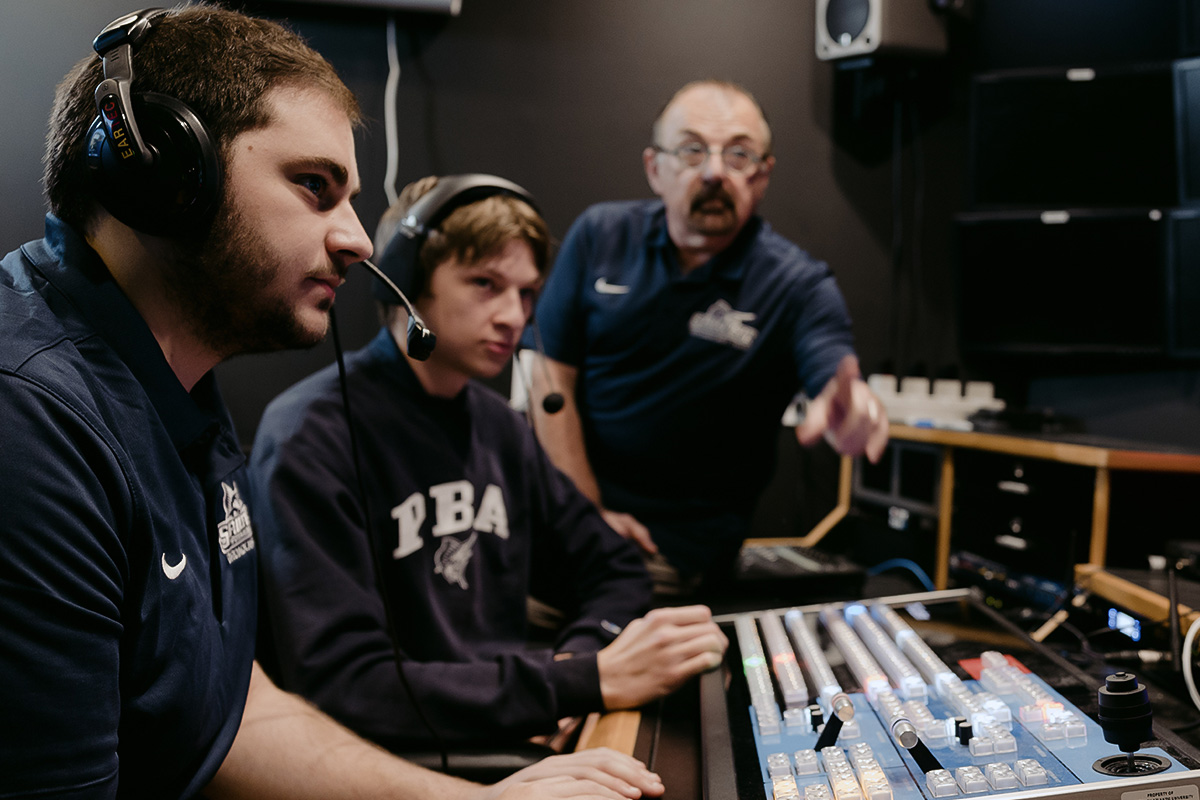 Male sports broadcasters wearing headsets and operating a professional mixing console in a dark television production studio
